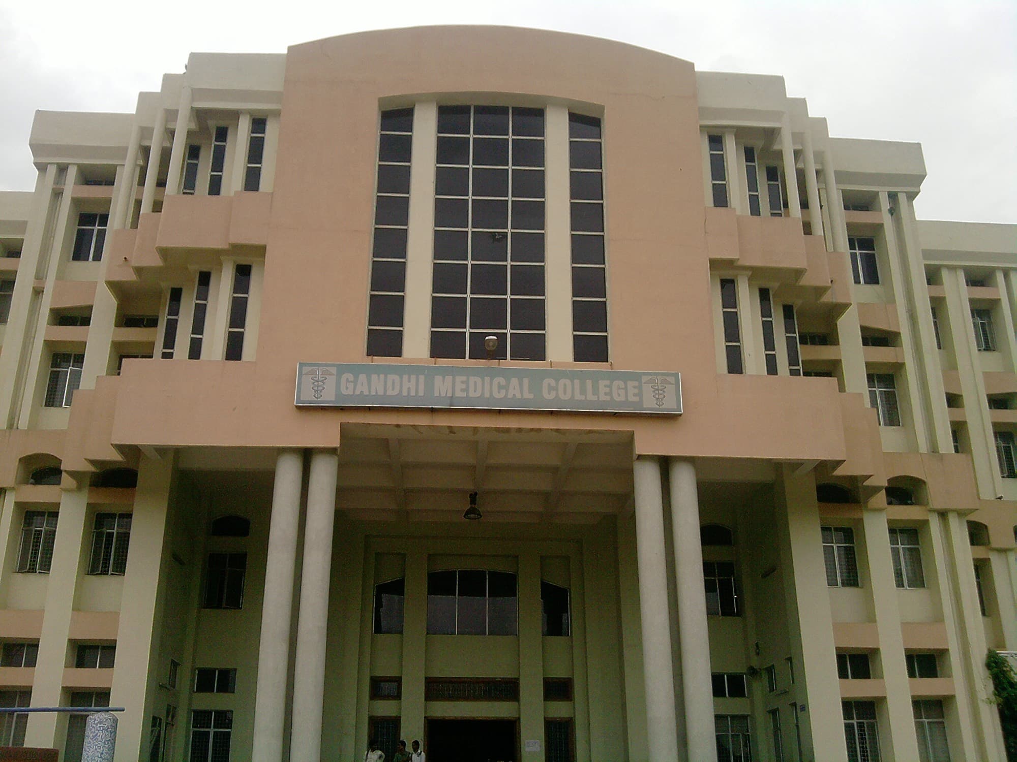The peach and cream facade of Gandhi Medical College, its name on a sign above a pillared porch, seen from close in on an overcast day.