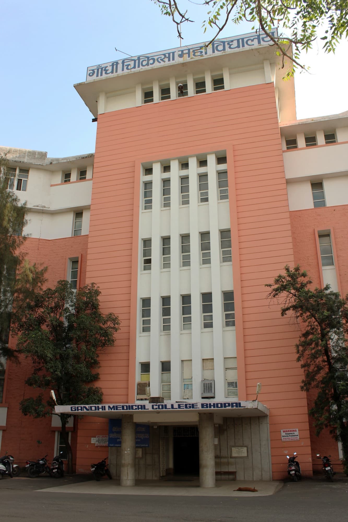 The salmon-pink central tower of Gandhi Medical College, Bhopal, with its Hindi name on the roof and 'Gandhi Medical College, Bhopal' over the entrance, motorbikes parked in front.
