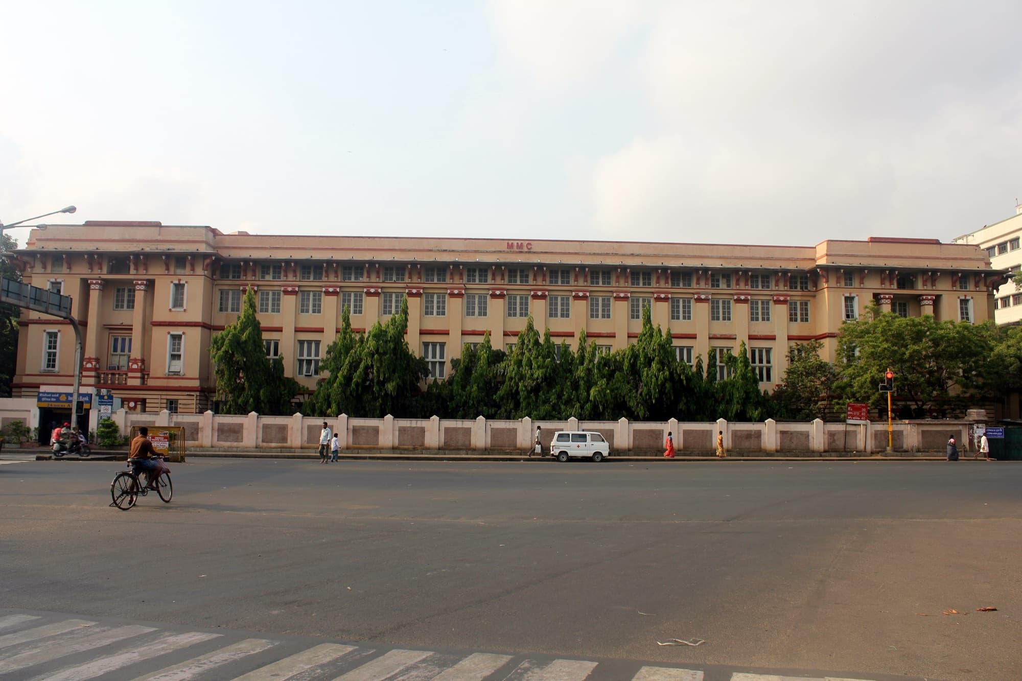 The long cream and maroon three-storey front of Madras Medical College, with 'MMC' on the parapet, behind a boundary wall and trees across an empty road.