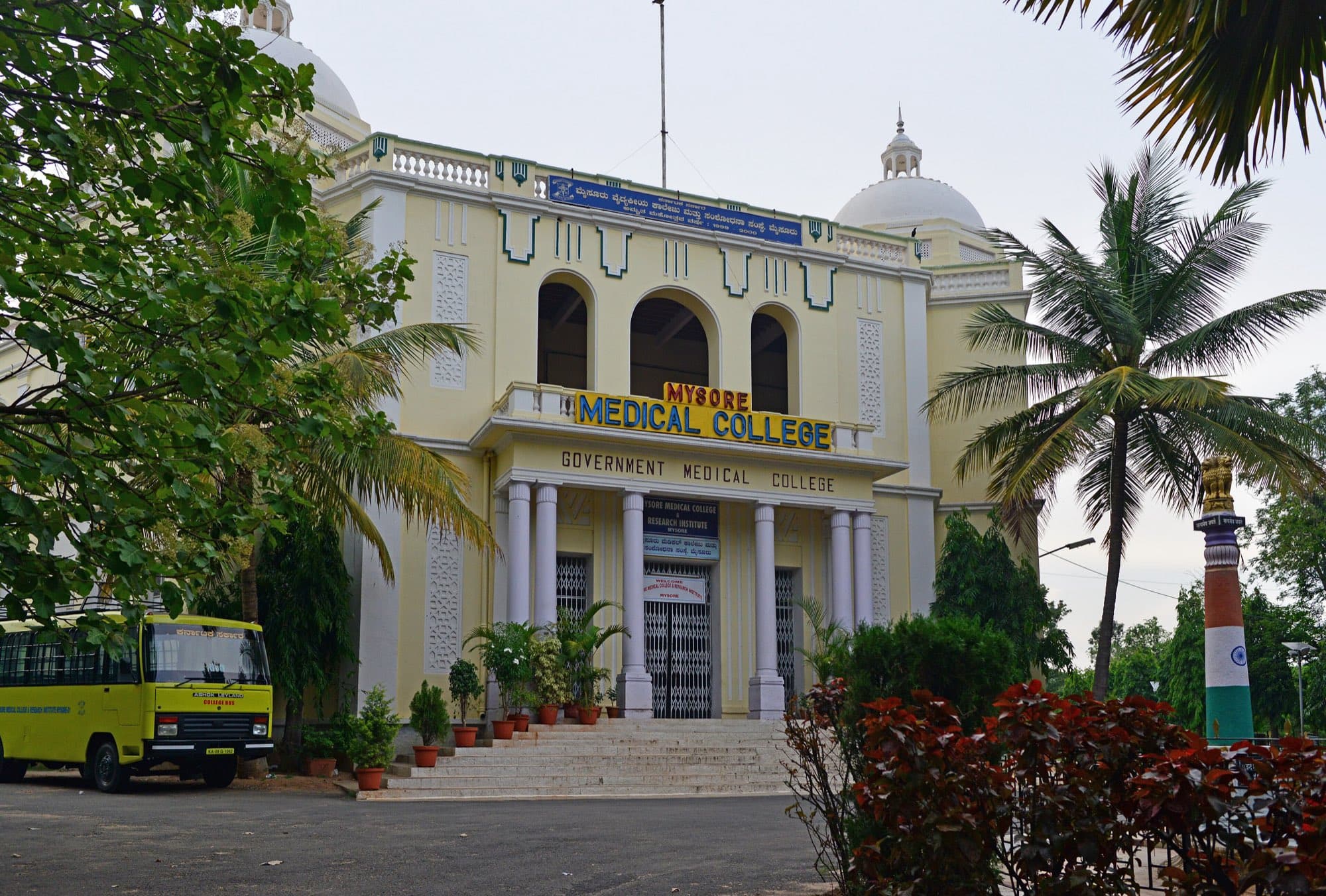 The pale yellow domed front of Mysore Medical College with 'Mysore Medical College' and 'Government Medical College' over a columned porch, a yellow college bus on the left and palm trees.