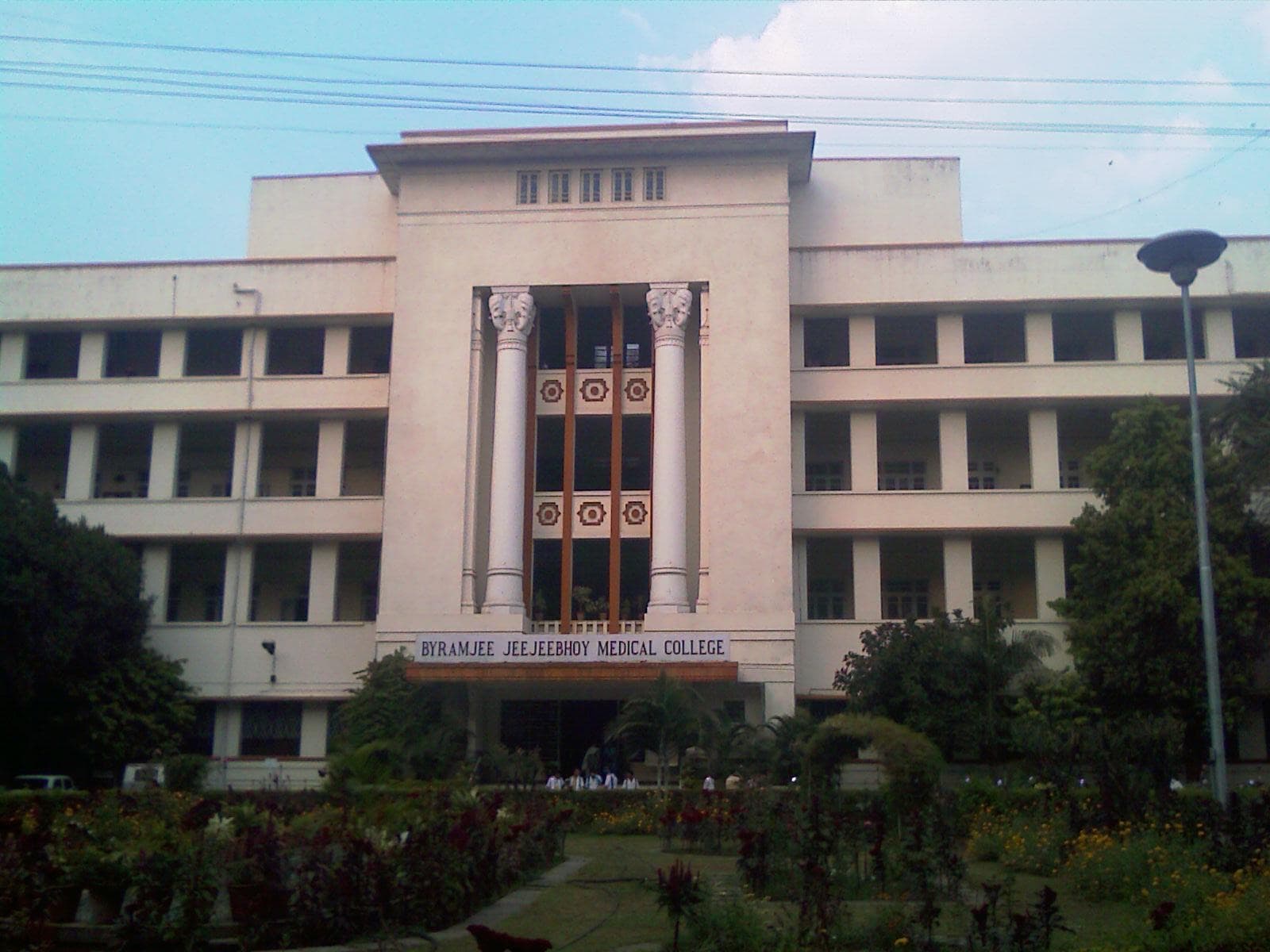 The cream-coloured four-storey front of Byramjee Jeejeebhoy Medical College, its name in large letters over the entrance, behind a flowering garden under a blue sky.