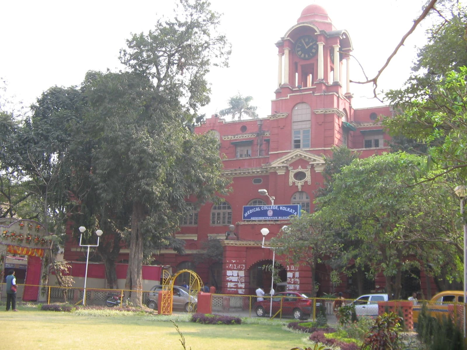 The red Administrative Block of Medical College, Kolkata, with its domed clock tower and a blue 'Medical College, Kolkata' sign, framed by trees and a lawn in hazy daylight.