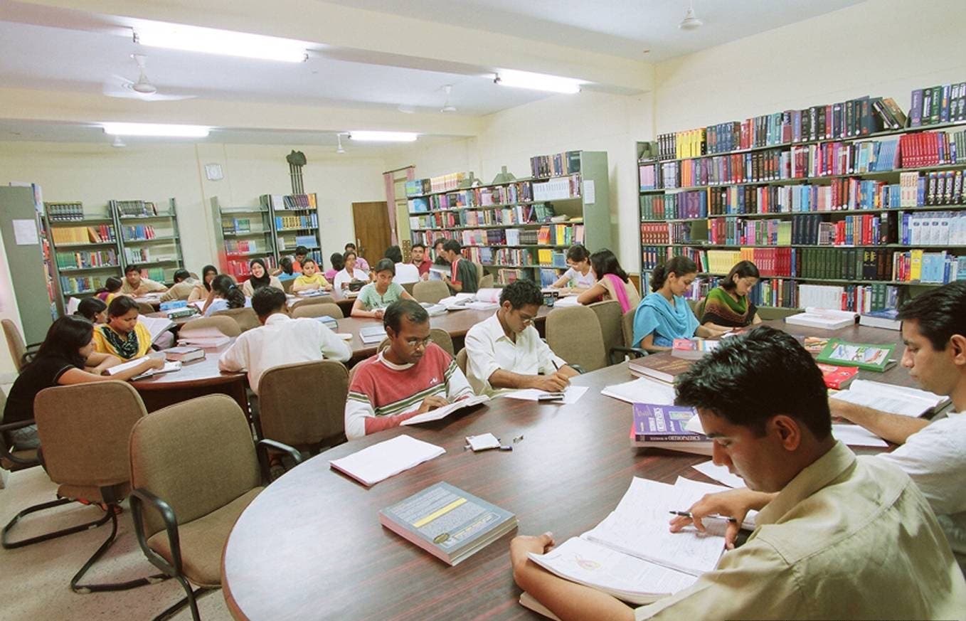 Medical students reading textbooks at shared tables in a college library, shelves of clinical volumes behind them.
